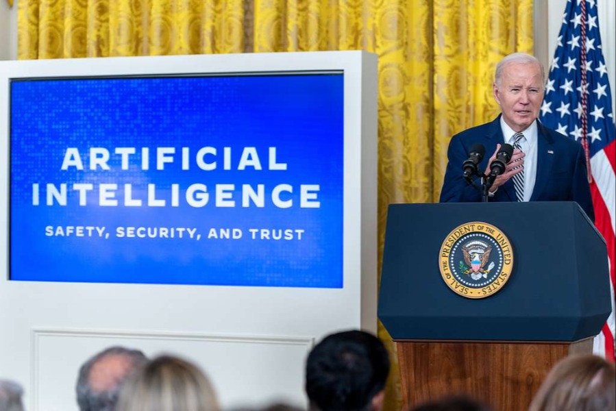 Image shows  President Joe Biden delivering remarks behind a podium, next to a blue screen displaying the words 