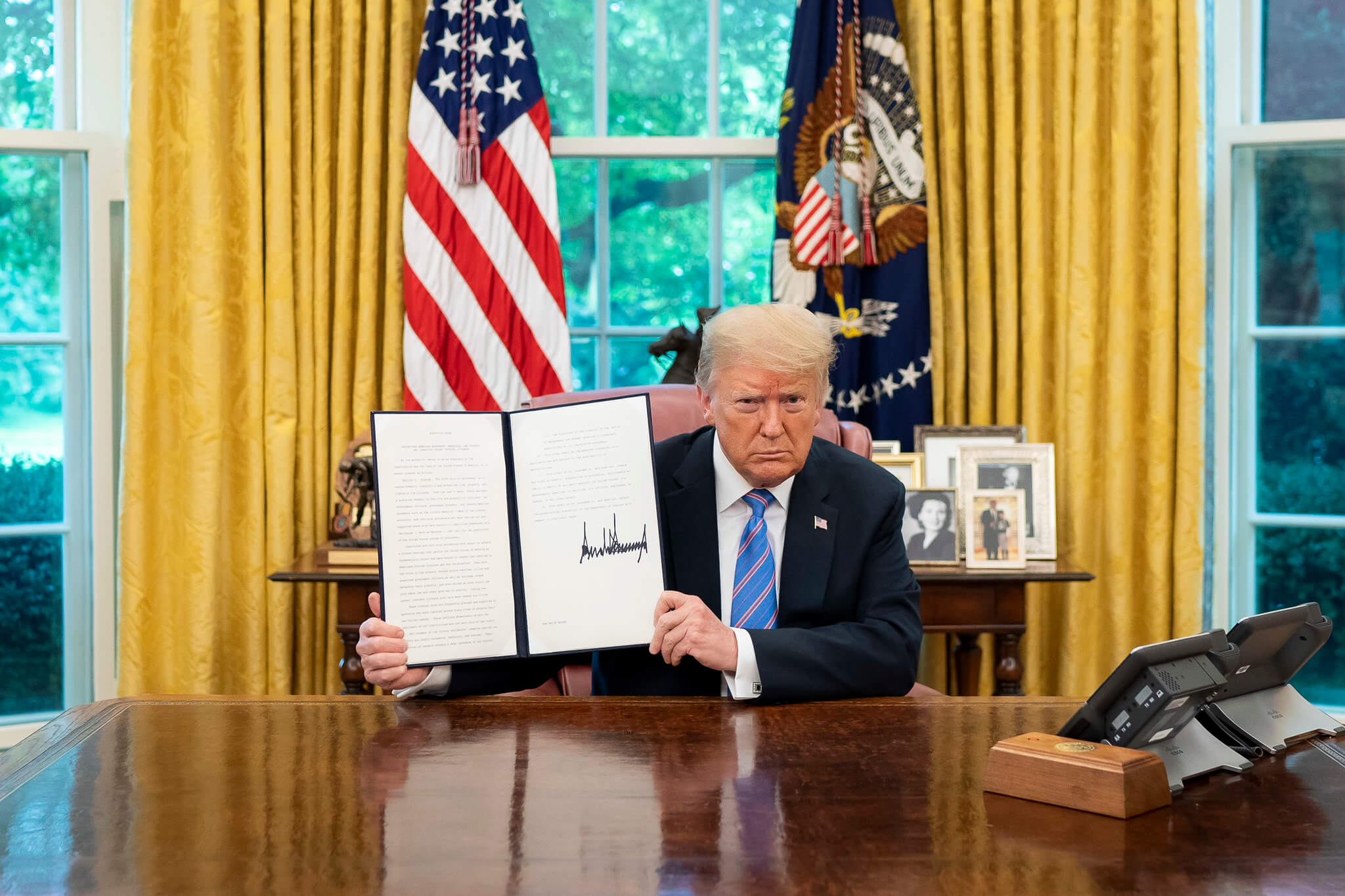 Trump holding an executive order sitting at his desk in the Oval Office